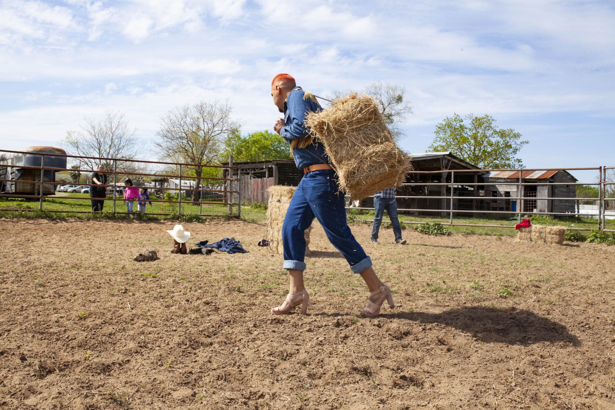 Jose Villalobos' installation A Las Escondidas (2019). The photo shows the artist carrying bales of hay on his back while wearing high heels.