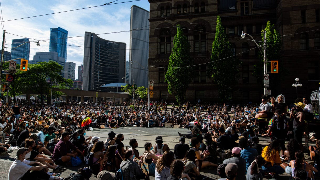 A crowd seated in a circle listen to speakers.