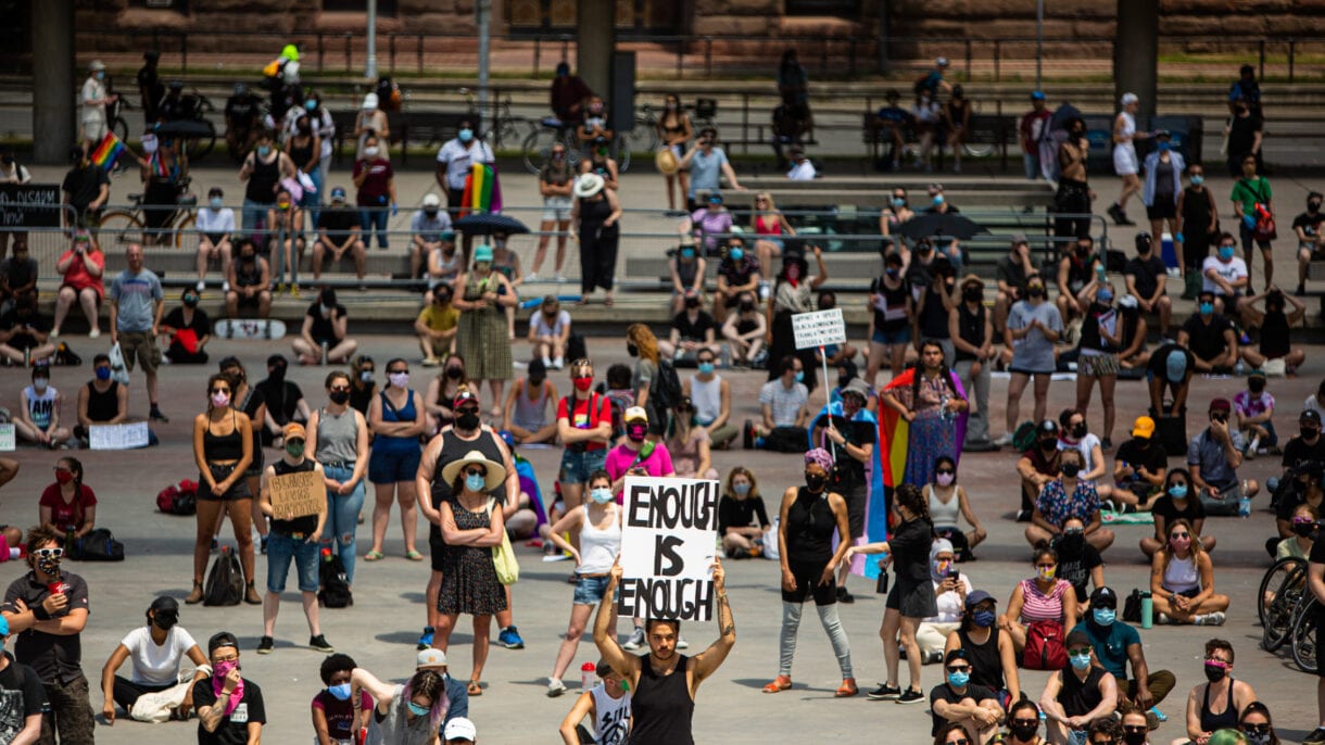 Among a crowd of protesters keeping a lot of space between them, one person is seen holding up a sign stating, 