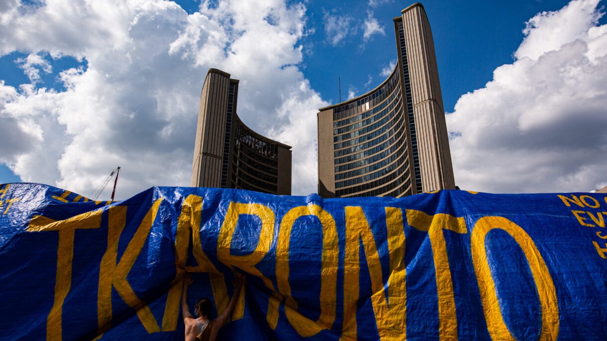 Activists hoist a giant tarp with the word Tkaronto on it over the Toronto sign in front of City Hall.