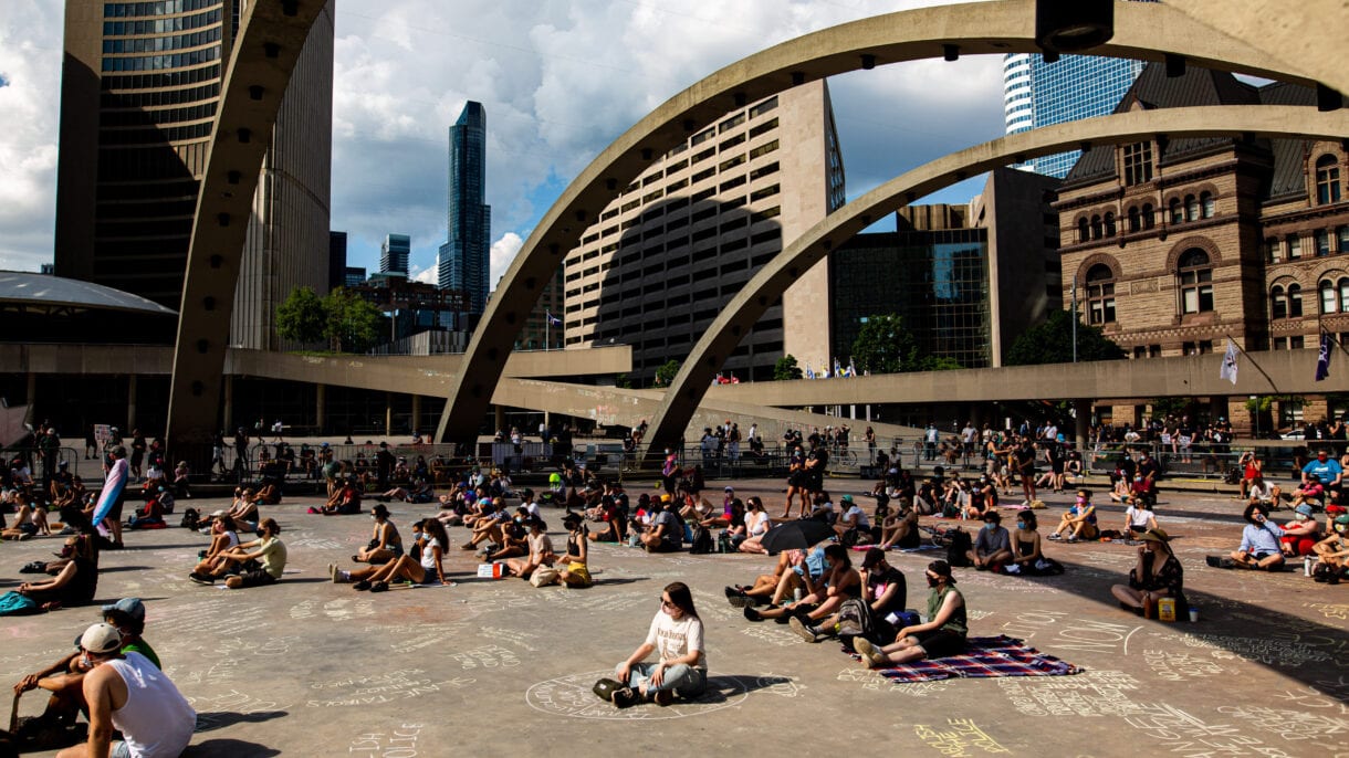 People sit in small groups in Nathan Phillips Square with the arches from the fountain and Old City Hall visible.
