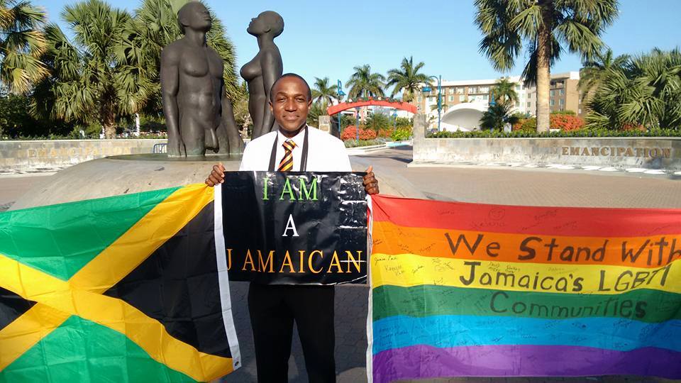 Maurice Tomlinson poses with a sign, Pride flag and Jamaican flag.