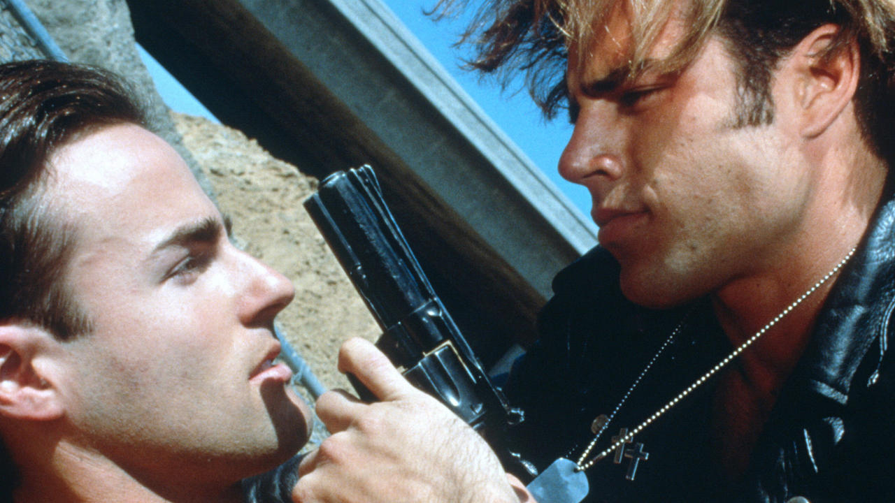 A man in leather holds up a gun to the face of another man.