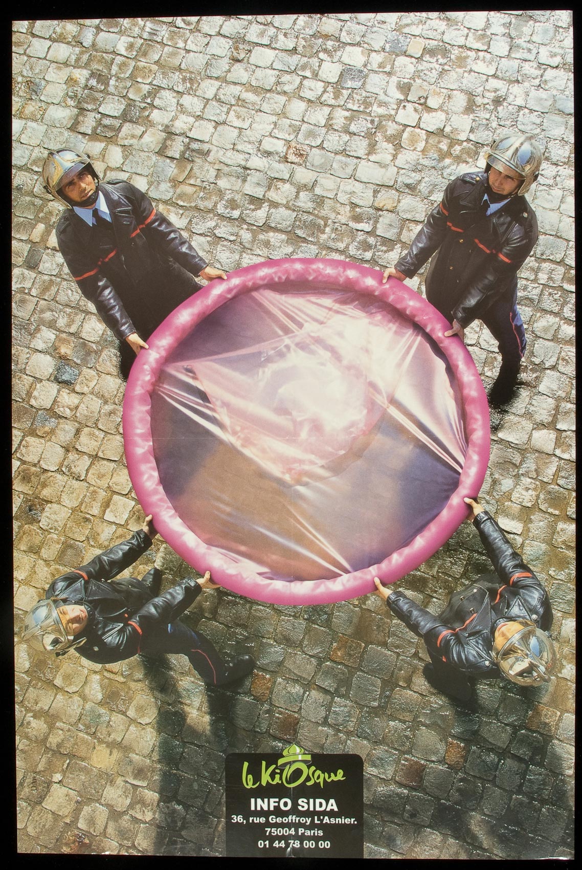 Four fireman seen from above hold an oversized pink condom still rolled up in a circle, as if it's a safety net ready for someone to jump into.