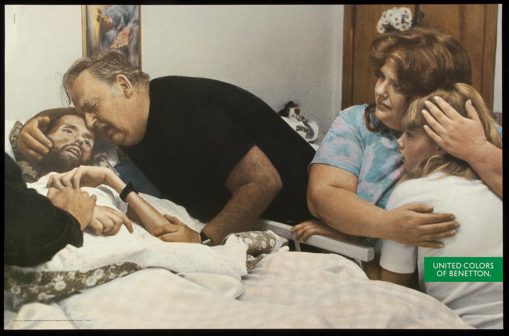 A frail emaciated young man is cradled by his crying father has his mother and sister look on distraught. From out of frame another hand emerges to hold the young man's hand.