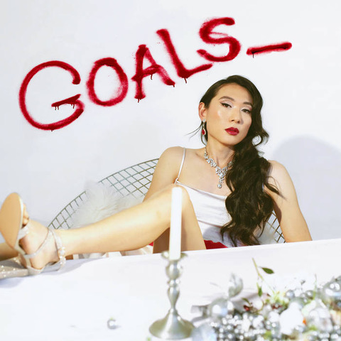 A woman leans back in a chair with her leg on the table in front of her and the word Goals_ scrawled on the wall behind her.