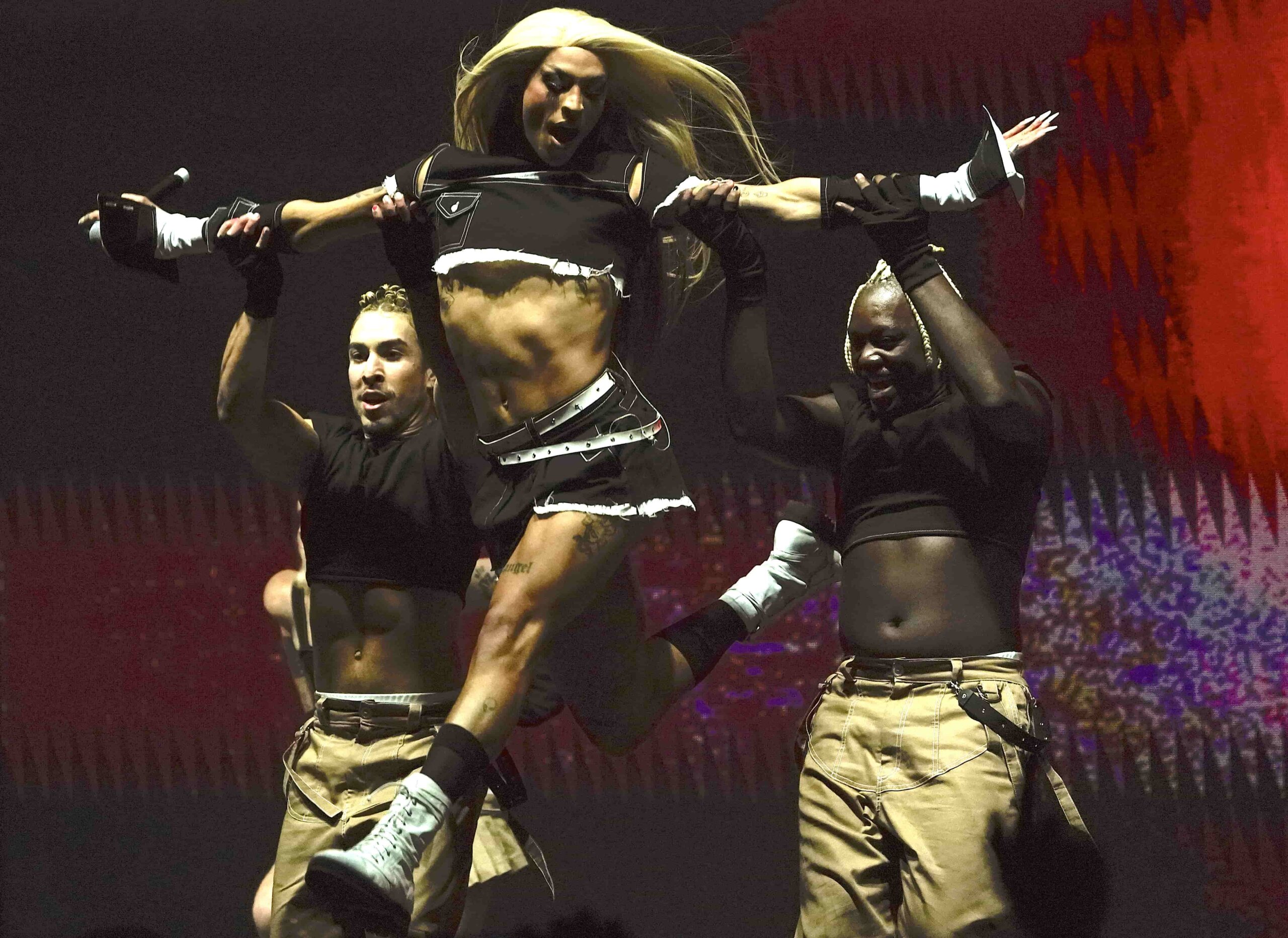 A fit young queen in a blonde wig is lifted up by two dancers during a concert performance.