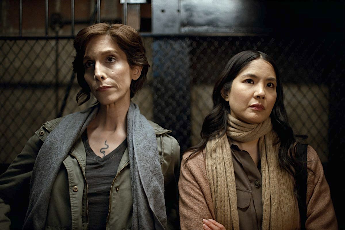 Two women lean against a chain link fence looking... perturbed.