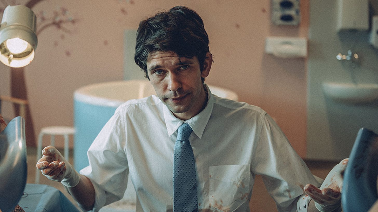 Ben Whishaw in a bloodied shirt and surgical gloves looks toward the camera from between the stirrups on an examination table.