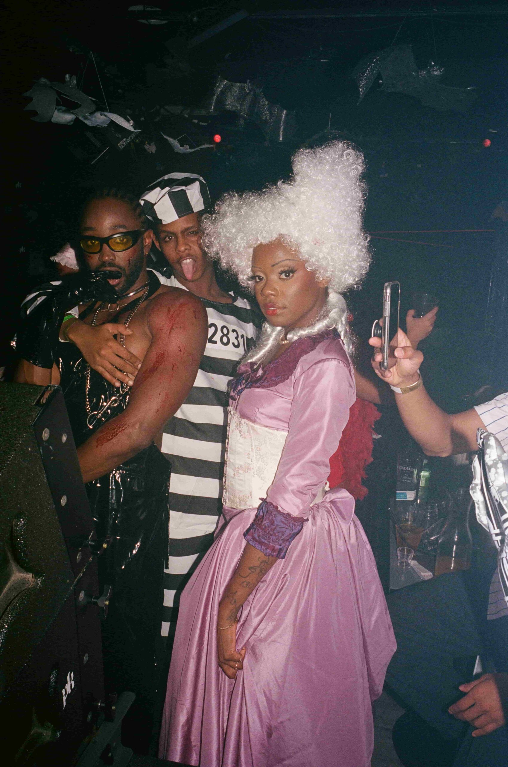 Two people of colour in costumes, one a pink 18th century gown and white wige, the other in black PVC, stand among party goers.