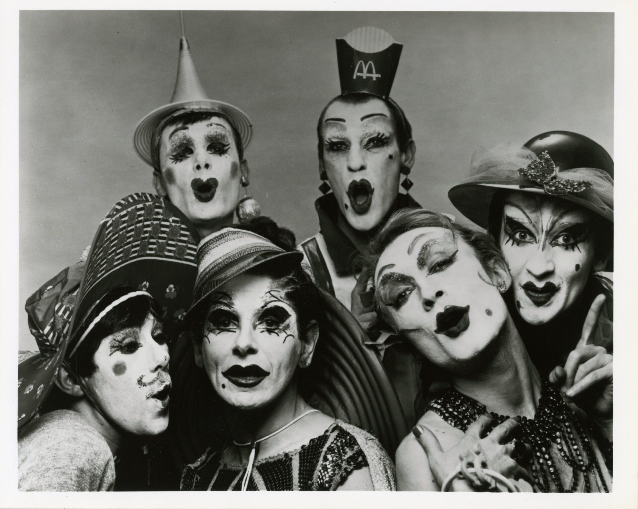 Black and white photo of six drag queens in old timey make-up; most wear hats, one has a McDonald's fry container on her head