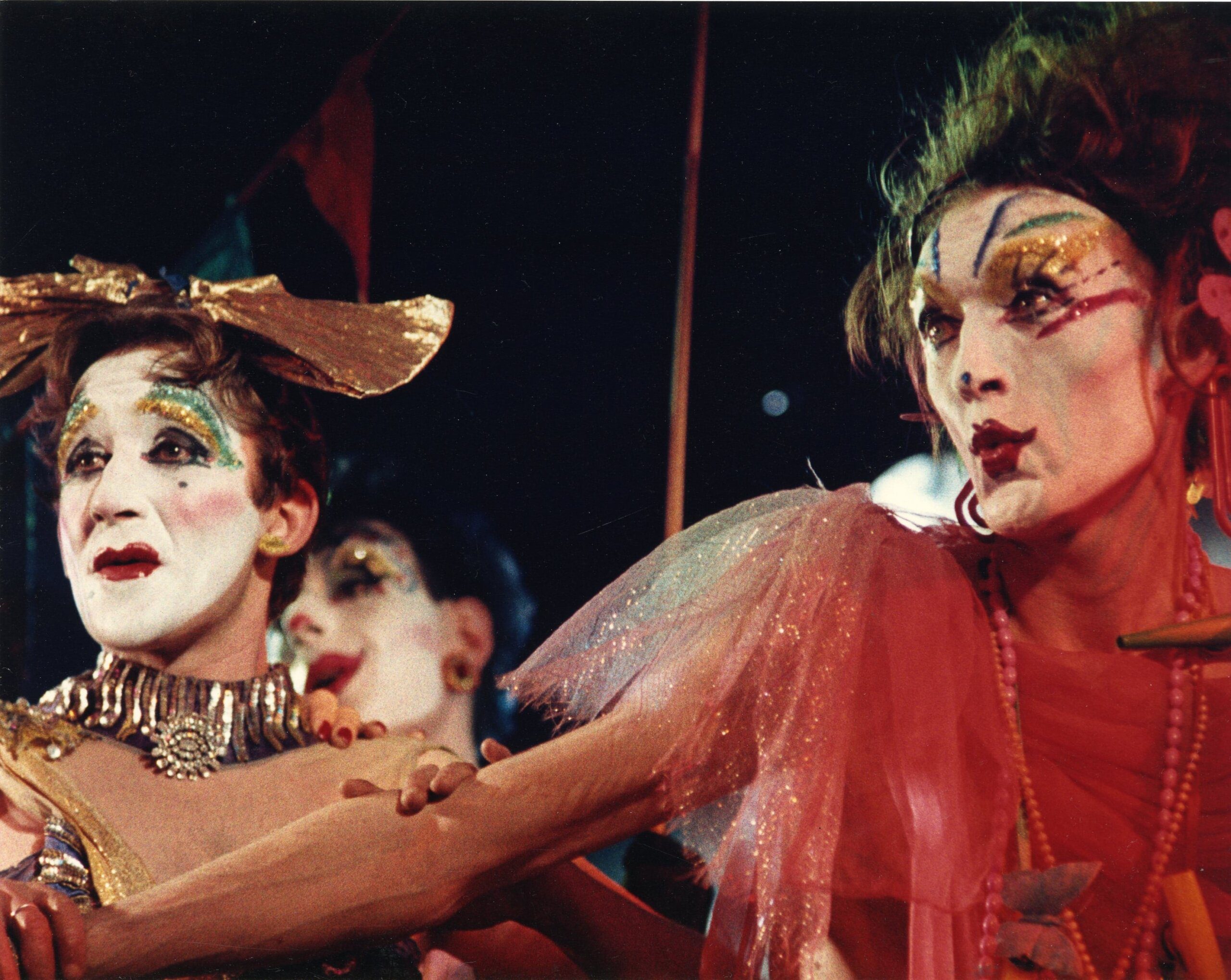 Colour archival image of two queens in glittery make-up; one wears gold lamé, the other a sheer pink shimmery fabric
