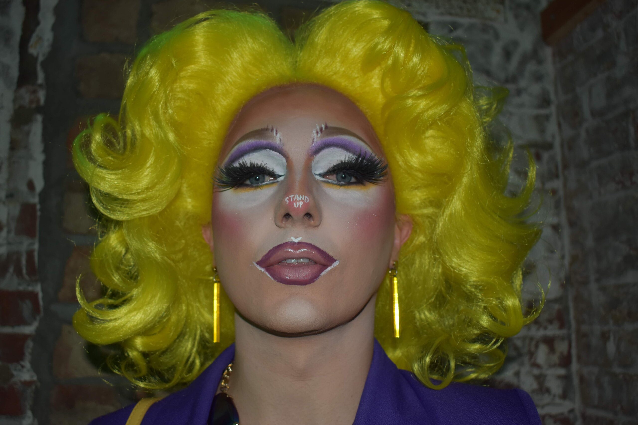 Drag queen Robin Rose Quartz wears a yellow wig and dramatic makeup; she looks directly at the camera.