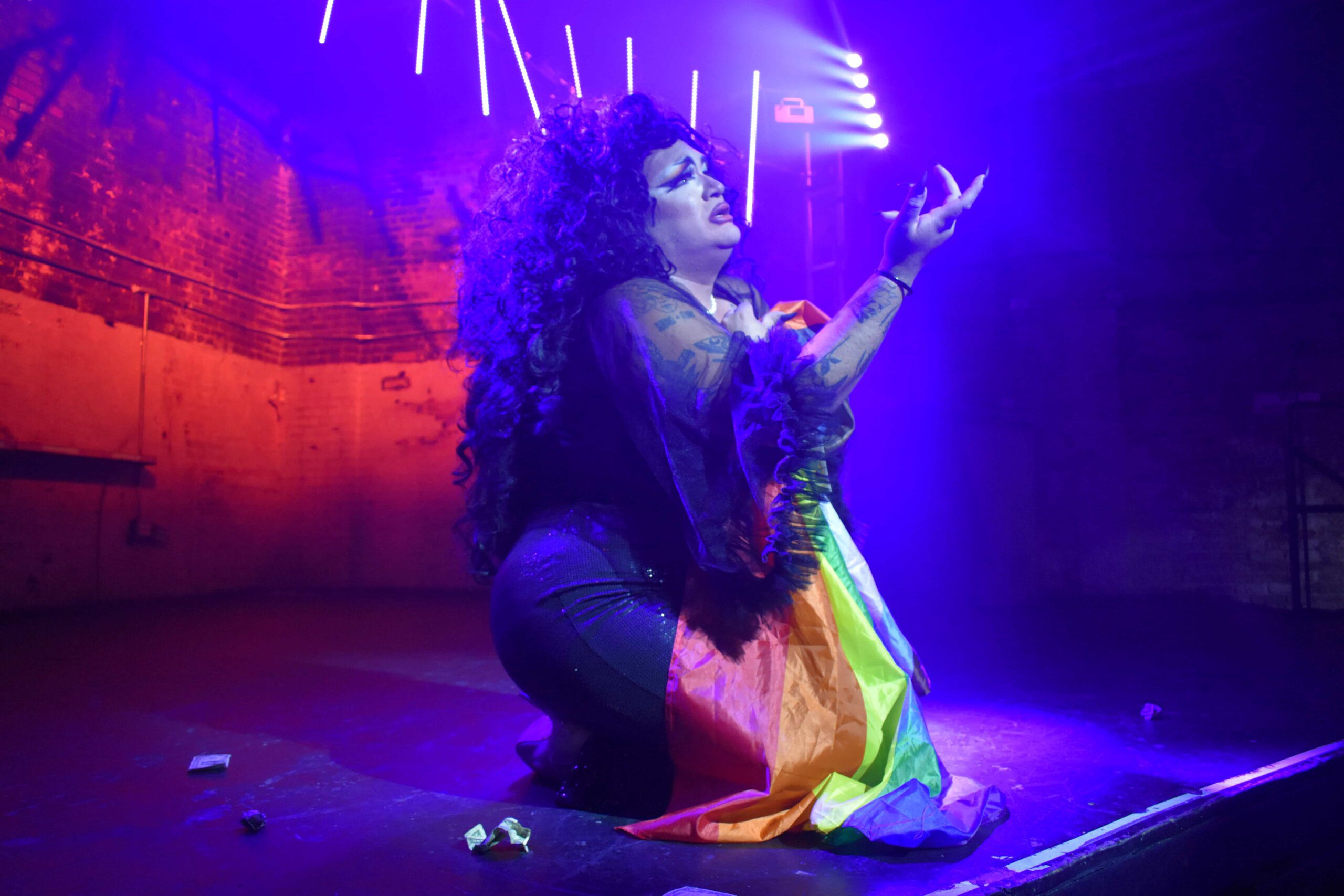 Drag queen Megami kneels on the stage in a long black curly wig and black sheer top; she holds the Pride flag to her chest.