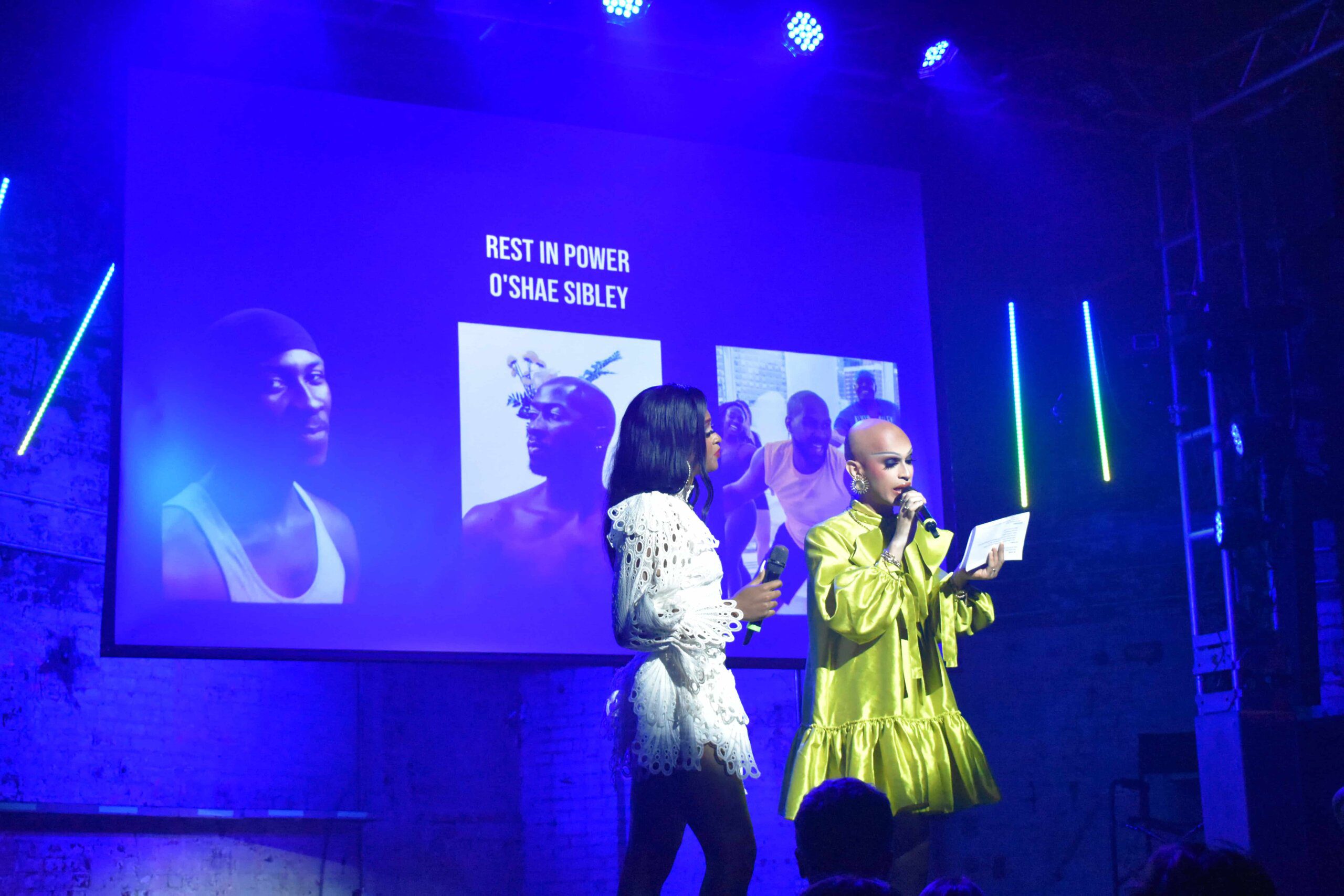 Two hosts, one in a greenish yellow dress and one in a white lace dress, hold microphones. The one in yellow reads from a folded paper. On a screen behind them are the words "REST IN POWER O'SHEA SIBLEY" and three photos of Sibley.