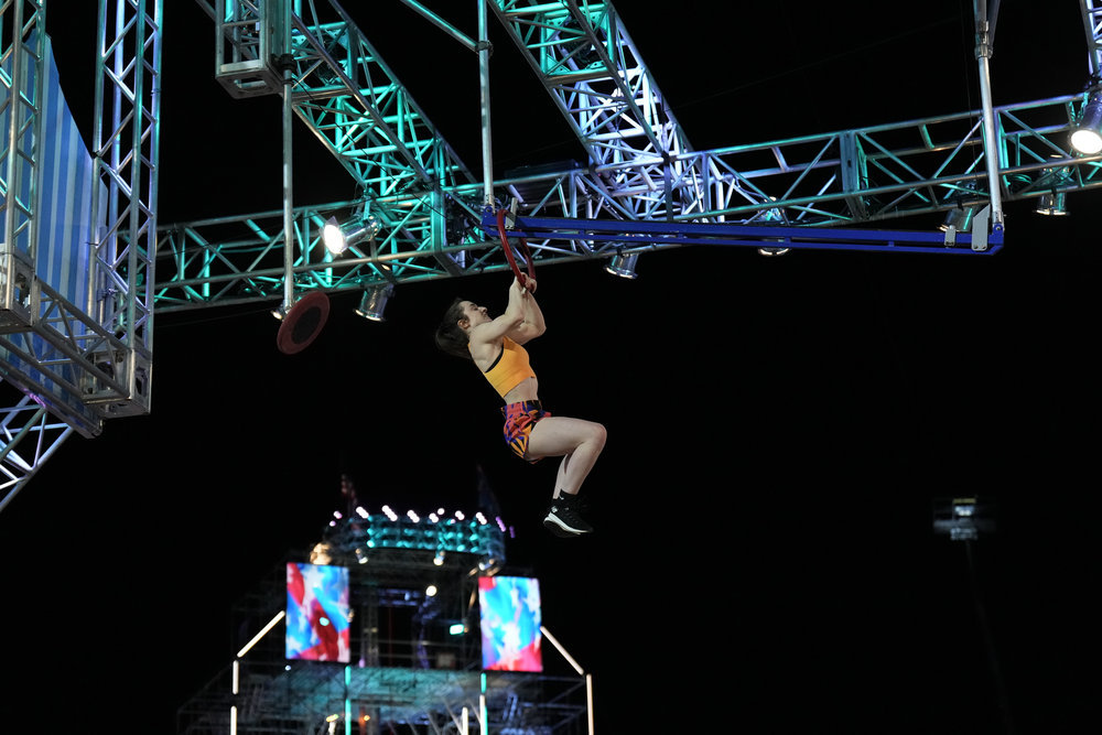 Casey Rothschild wears orange, pink and black. She holds herself up while hanging from a rafter.