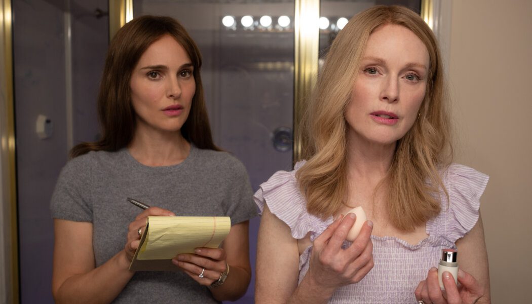 Natalie Portman as Elizabeth Berry and Julianne Moore as Gracie Atherton-Yoo in May December. Portman wears a grey t-shirt and holds a pen and yellow pad of paper; Moore wears a top with ruffles on the shoulders.