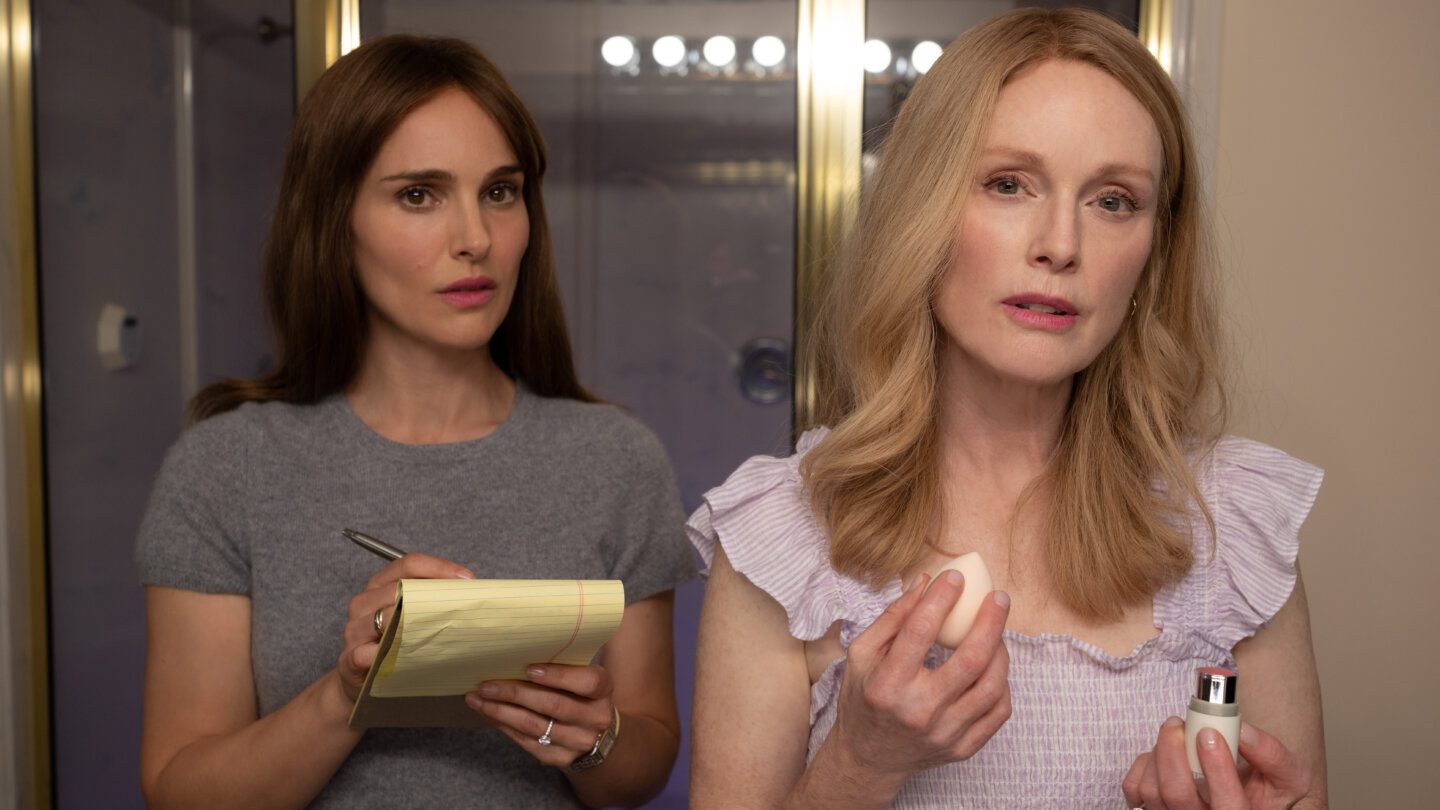 Natalie Portman as Elizabeth Berry and Julianne Moore as Gracie Atherton-Yoo in May December. Portman wears a grey t-shirt and holds a pen and yellow pad of paper; Moore wears a top with ruffles on the shoulders.