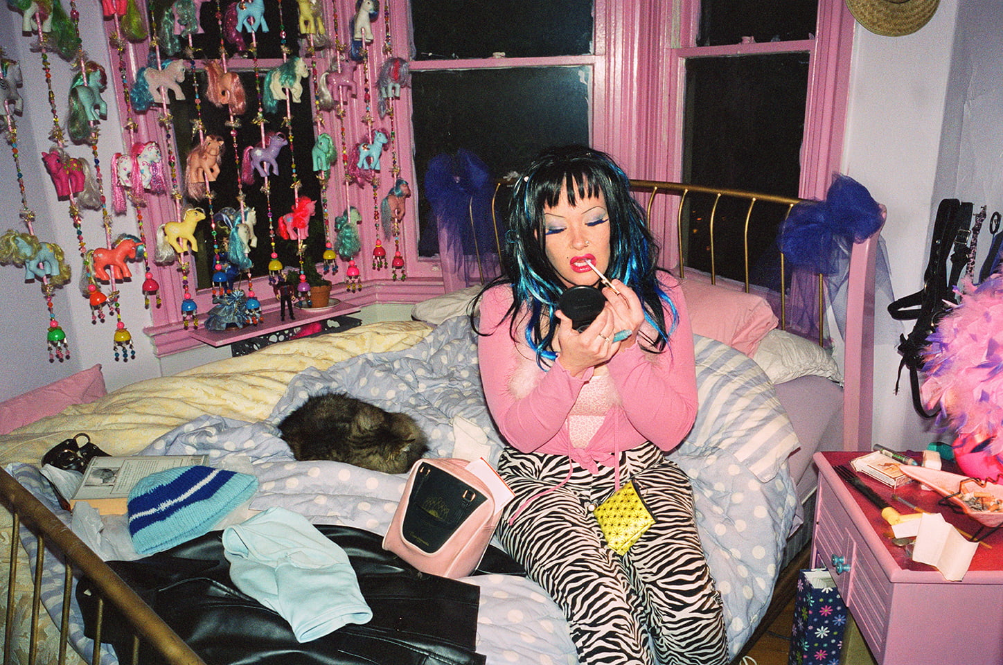 A person with blue hair, a pink top an zebra print pants sits in a busy bedroom. The window frames are painted pink and a beaded curtain incorporating My Little Pony toys hangs behind them.
