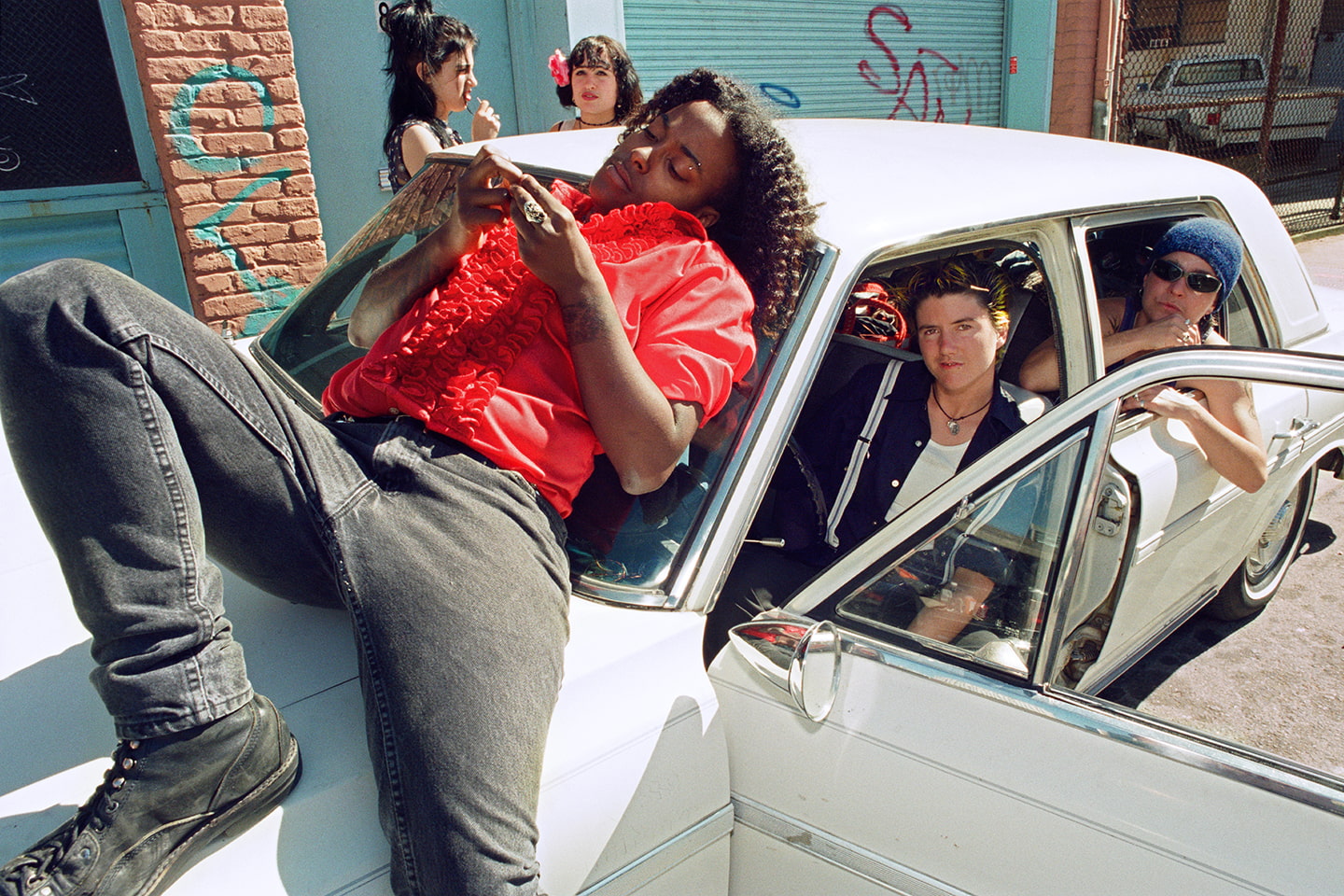 Five people in and around a white car. One person lays on top of the hood; they wear a red tuxedo shirt with ruffles.