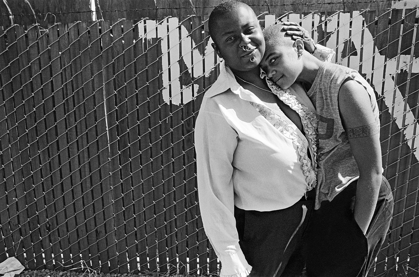 Two people with shaved heads stand in front of a chain link fence. One leans on the other, who cradles their head.
