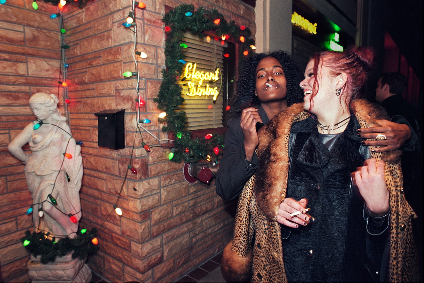 Two people smoke cigarettes in front of Christmas lights and a neon sign that says "Elegant Dining." One wears an open leopard print coat.