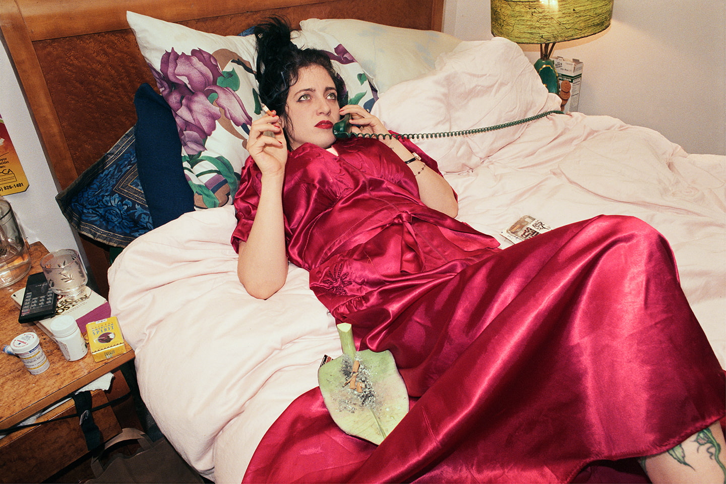 A person with dark hair holds a cigarette and a phone with a spiral cord. They wear a red satin dress or robe and have a leaf shaped ash tray. They are laying on a white bed.