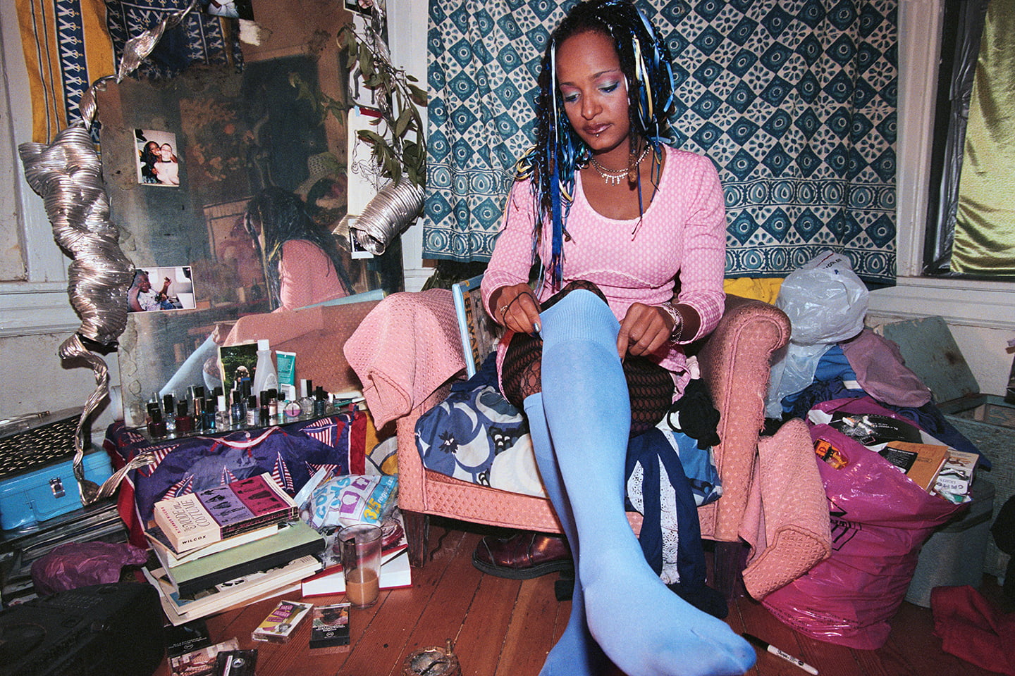 A person in pink and blue with long hair pulls up a knee-high in a messy bedroom