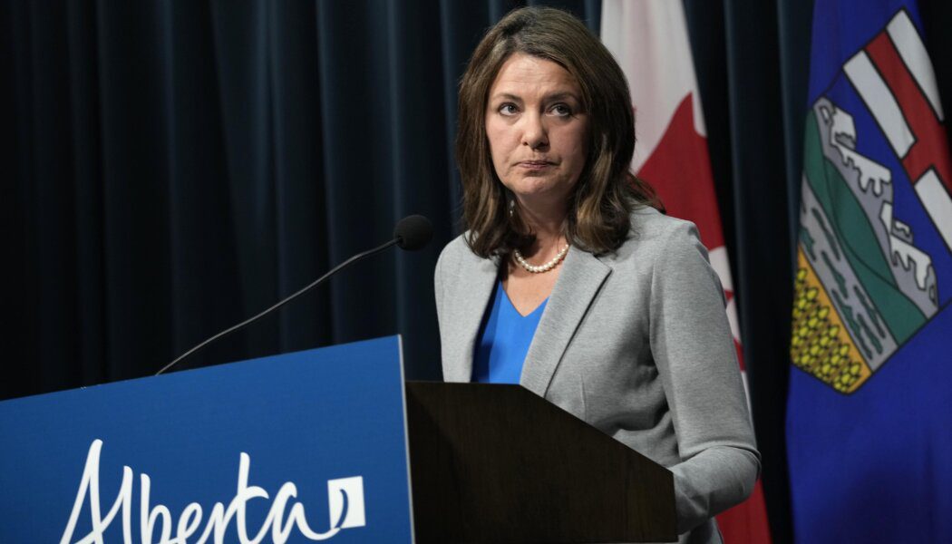 Danielle Smith wears a blue top, grey blazer and pearls. She stands behind a podium with an Alberta sign, in front of Canadian and Alberta flags.