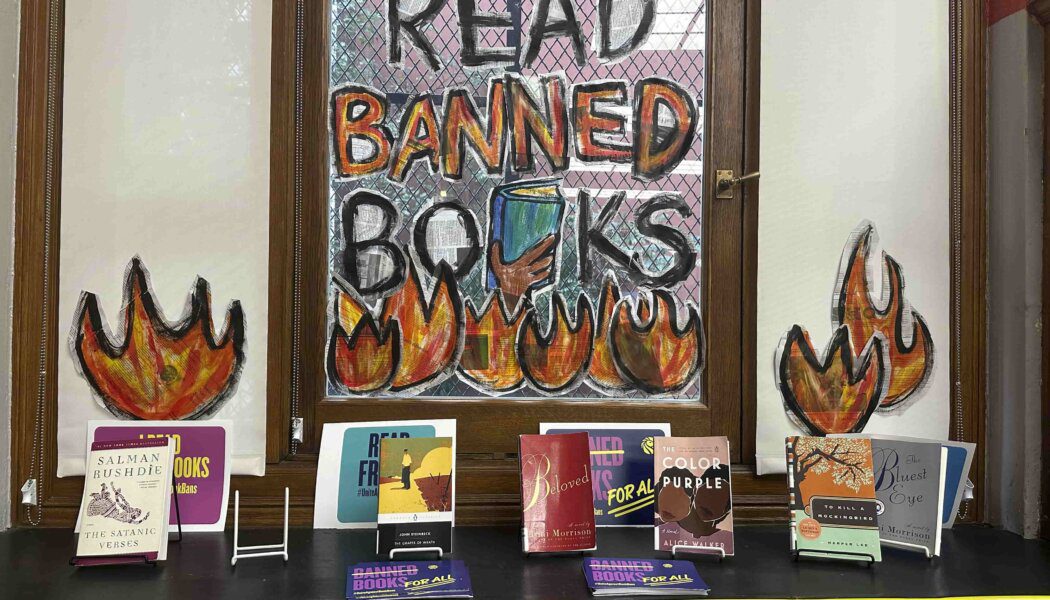 "Read Banned Books" is written on a window above flames; below, an assortment of banned books are lined up above Caution tape.