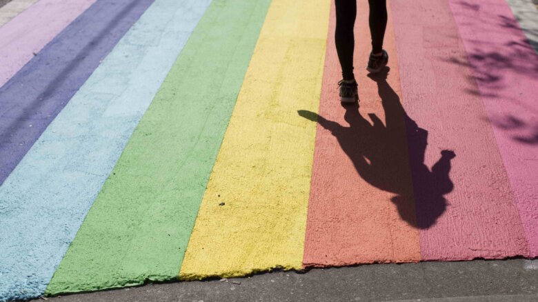 A person's legs and feet are seen on a rainbow crosswalk; their shadow is visible.
