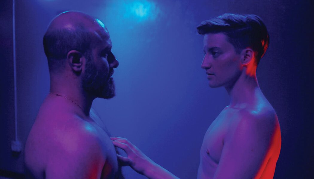 Theo Germaine and Aden Hakimi are lit in purple; they are both shown from the chest up, shirtless. Germaine touches Hakimi's chest while the pair face each other. Hakimi is balding and has a short beard; Germaine has short brown hair.