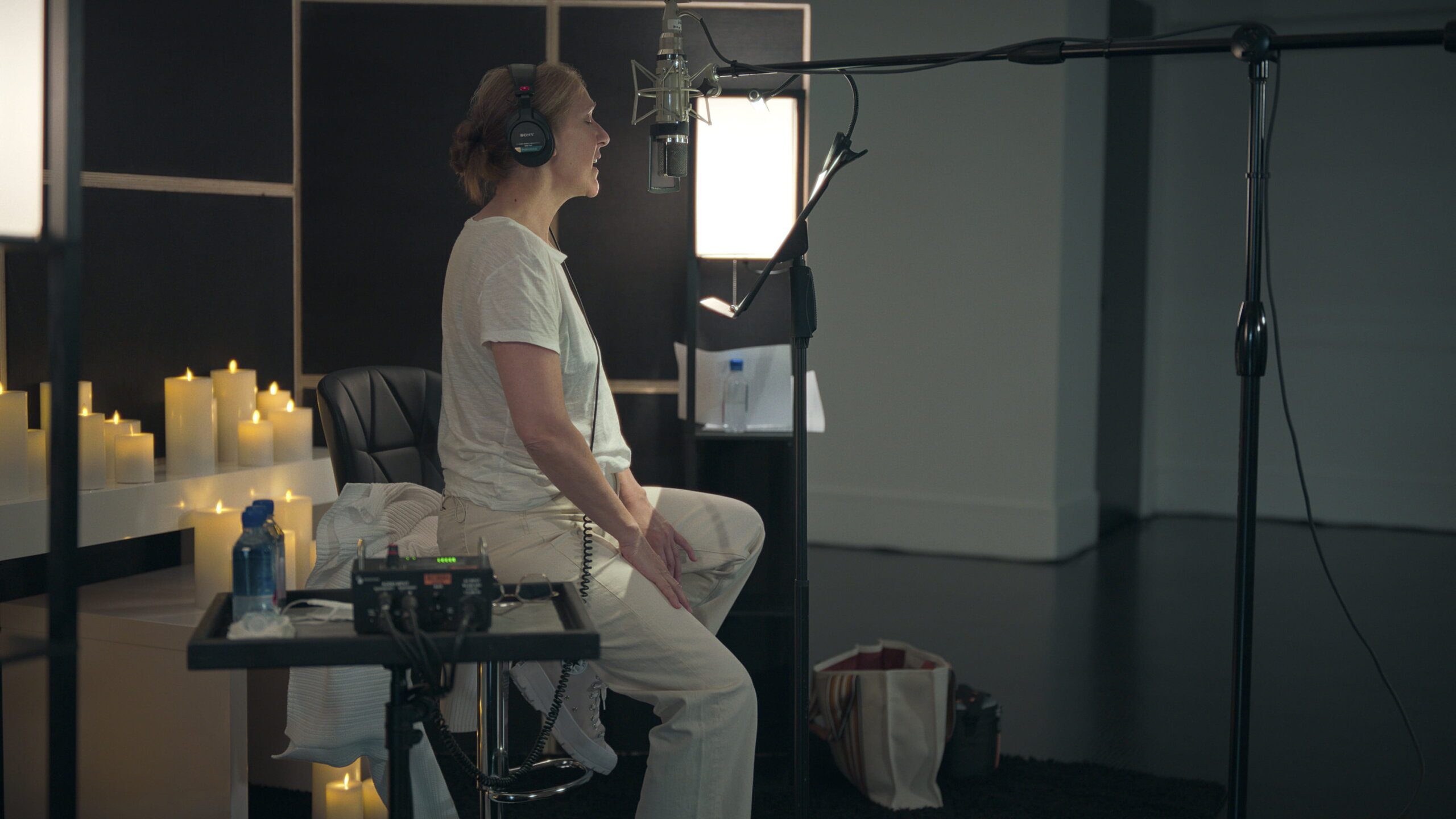 Celine Dion wears white and headphones; she sits in front of a microphone, singing. Behind her are white candles.