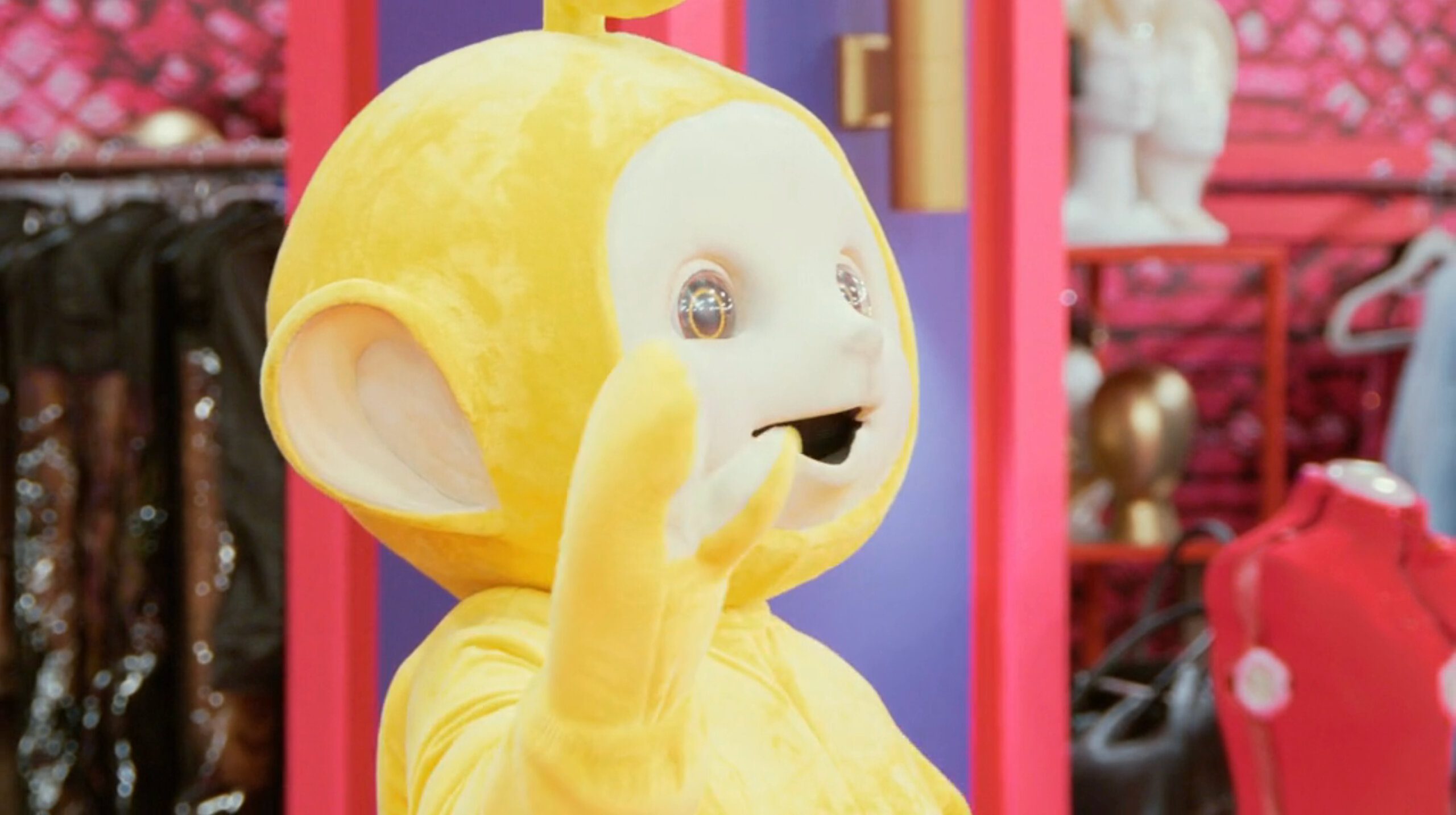 A yellow teletubby character with large ears and sparkling eyes waving, set against a colorful backdrop with clothing and mannequins in a scene from RuPaul's Drag Race All-Stars Season 9 Episode 5.