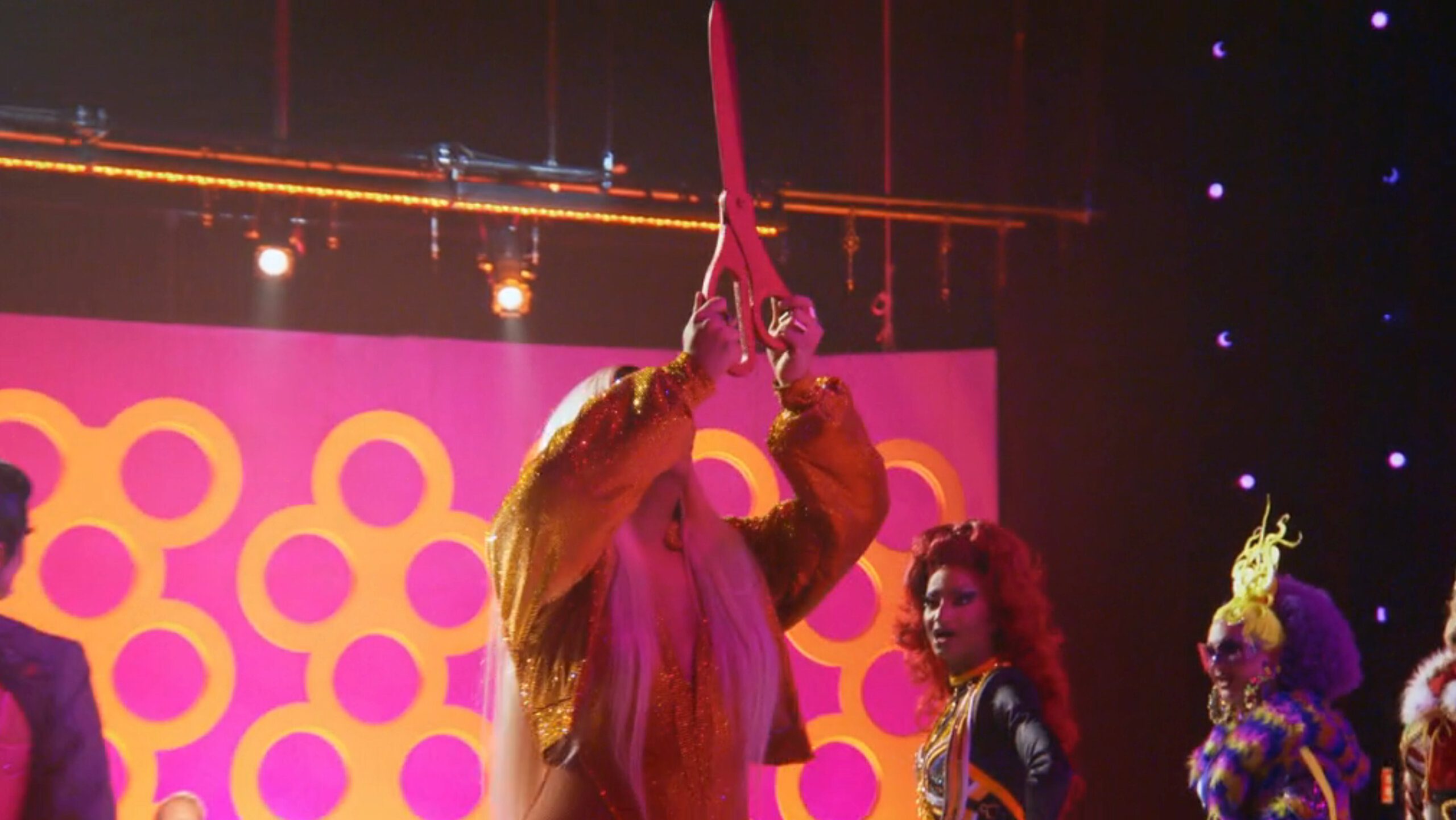 A performer in a glittering orange outfit holding oversized scissors above their head, surrounded by other vibrantly dressed individuals on stage with a pink background in a scene from RuPaul's Drag Race All-Stars Season 9 Episode 5.