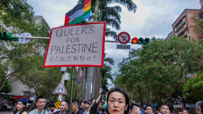 A person holds a placard reading "Queers for Palestine" during the Taiwan Pride parade on October 28, 2023 in Taipei, Taiwan. A Palestinian flag with a rainbow flag at one end is seen above.
