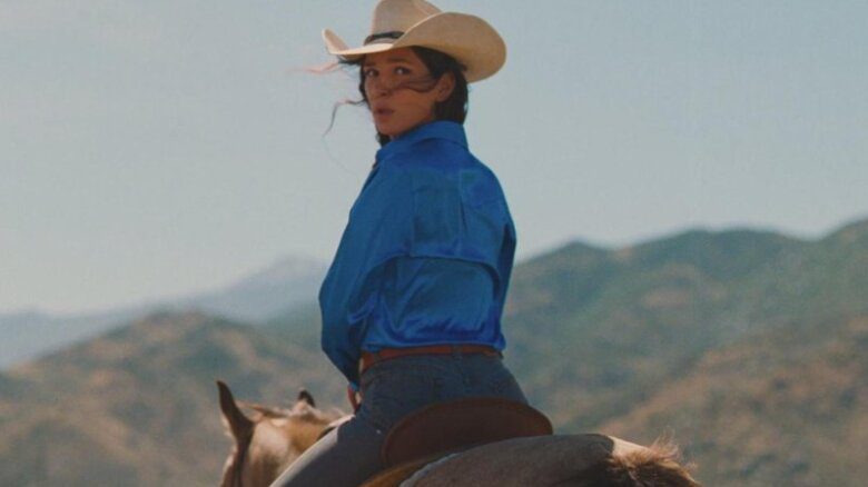 Eve Lindley from behind in a cowboy hat, blue button up, jeans and a brown leather belt riding a horse. She has long brown hair and looks over her shoulder.