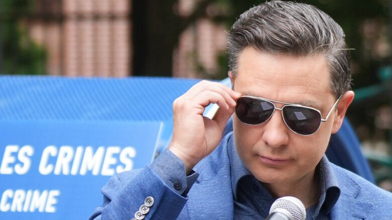 Federal Conservative Leader Pierre Poilievre arrives at a press conference in Montreal. He is outdoors, in front of a blue and white sign with its text cut off. He is shown from the shoulders up, wearing a blue suit and adjusting his aviator-style sunglasses. A microphone is in front of him.