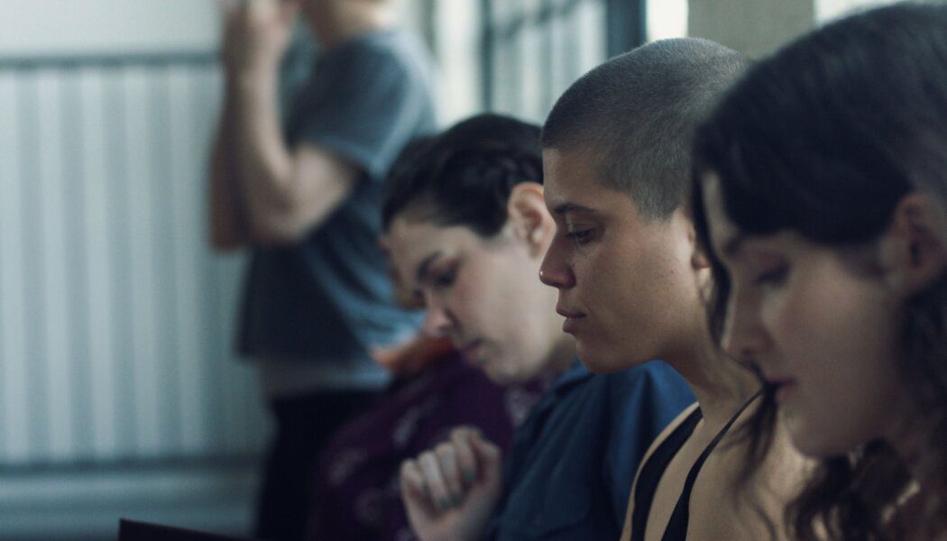 Breton Lalama in a row of others in a still from Really Happy Someday. Lalama has a shaved head is seen from the shoulders up in profile between two people with and brown hair.