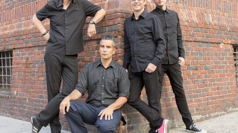 All four members of Pansy Division wear black pants and black button-up shirts. They are shown leaning against the corner of a red brick wall.