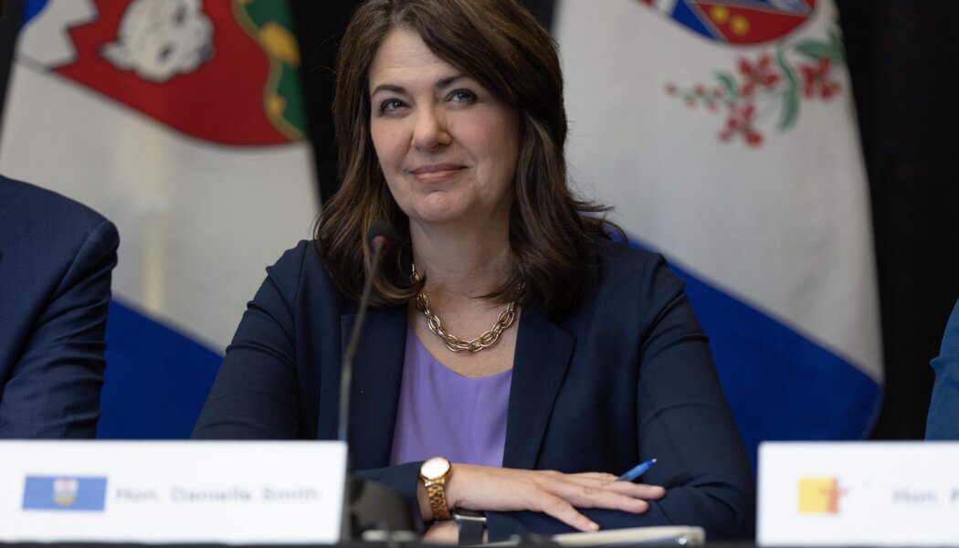 Alberta Premier Danielle Smith sits behind a microphone; she wears a blazer, purple top and gold necklace and watch. Northwest Territories and Yukon flags are shown behind her.