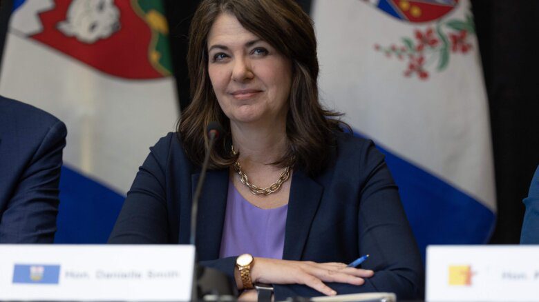 Alberta Premier Danielle Smith sits behind a microphone; she wears a blazer, purple top and gold necklace and watch. Northwest Territories and Yukon flags are shown behind her.