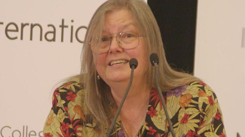 Dorothy Allison sits behind a microphone. She has long, light-coloured hair and wears glasses and a patterned button-up shirt.