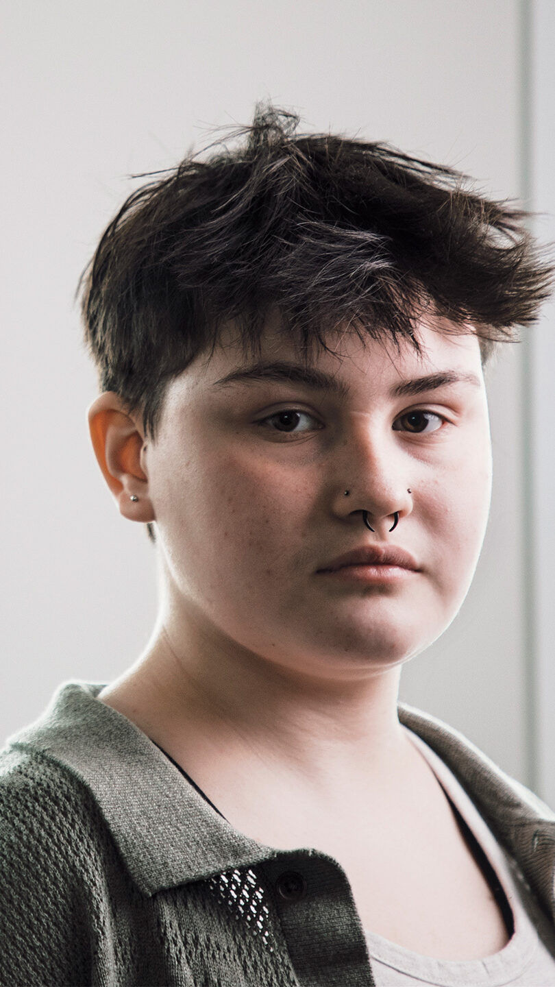 A young person with short dark hair and an olive green shirt faces the camera; a Monstera leaf is to one side of them. They have their nose and septum pierced.