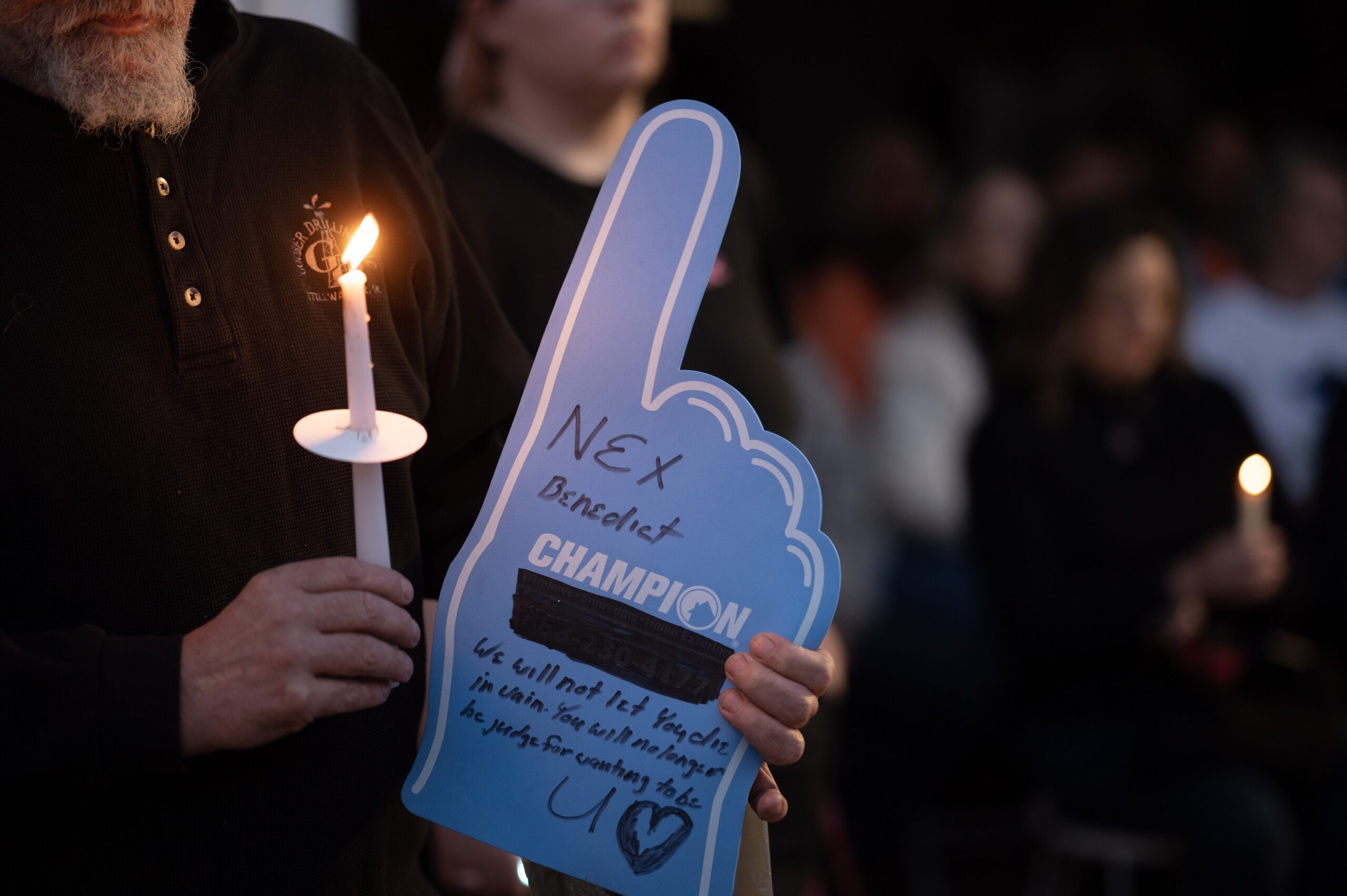 A person in black carries a candle and a sign that says "Nex Benedict - We will not let you die in vain. You will no longer be judged for wanting to be you."