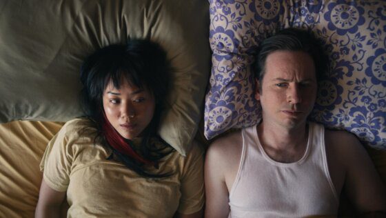 An Asian woman and a white man are side-by-side in a bed. She is on a green pillow; a yellow sheet visible below her. He is on a purple and white patterned pillow. She has a red streak in her hair and wears a light yellow t-shirt; he wears a white sleeveless undershirt.