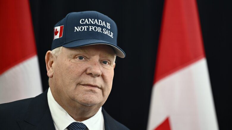 Ontario Premier Doug Ford, wearing a 'Canada Is Not For Sale' hat, arrives for a first ministers meeting in Ottawa on Jan. 15, 2025. Canadian flags hang behind him. He wears a suit and tie.