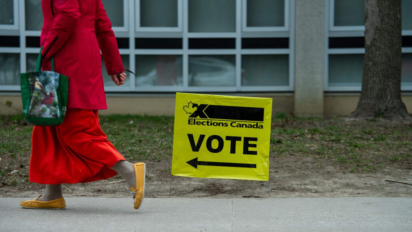 A person wearing all red clothes and yellow shoes carries a green bag; they walk past a yellow sign with black letters that reads "Elections Canada VOTE" with an arrow.