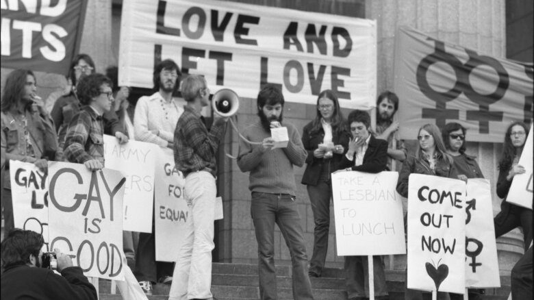 A black and white photo of speakers at a rally; a sign that says "Love and Let Love" hangs behind them