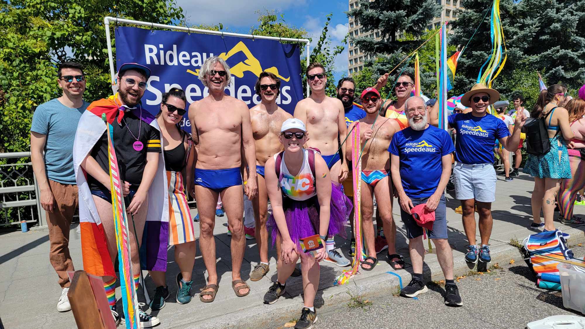 Members of Rideau Speedeaus gather before the Pride march, many in league shirts or swimsuits. They stand in front of a sign with the league's name on it.